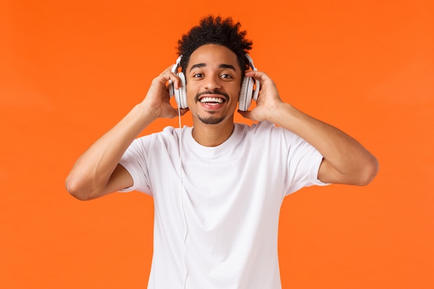 Conceito de felicidade, tecnologia e gadgets. homem afro-americano carismático feliz atraente em camiseta branca, ouvindo música em fones de ouvido, sorrindo câmera alegre, como presente, laranja