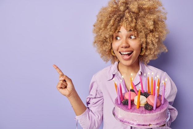 Conceito de feliz aniversário Mulher europeia alegre com cabelo loiro encaracolado segura bolo doce apetitoso com velas acesas aponta para longe no espaço da cópia usa camisa isolada sobre fundo roxo