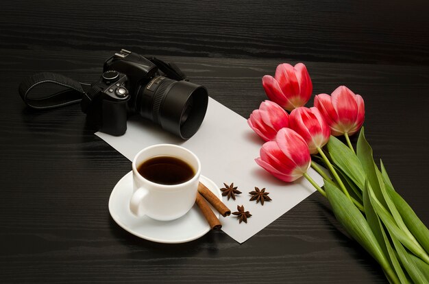 Conceito de férias. Buquê de tulipas cor de rosa, câmera dslr, xícara de café, canela, anis estrelado e folha de papel