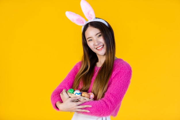 Conceito de férias da Páscoa, feliz jovem asiático com orelhas de coelho mão segurando uma cesta com ovos de Páscoa coloridos em isolado no fundo amarelo do estúdio do espaço da cópia em branco.