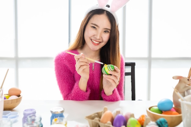 Conceito de férias da Páscoa, feliz mulher asiática com orelhas de coelho pintados à mão com ovos para a Páscoa com ovos de Páscoa coloridos no fundo do quarto branco