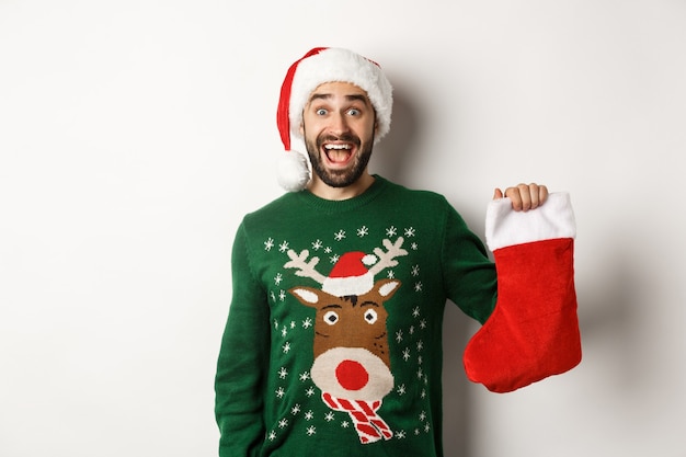 Conceito de férias de Natal e inverno. Homem feliz ganhou um presente na meia de Natal, parecendo animado, em pé com o chapéu de Papai Noel contra um fundo branco.