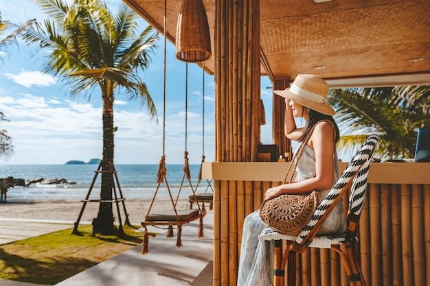 Conceito de férias de verão de viagens. Mulher asiática feliz viajante com chapéu e vestido relaxa em balanço no café da praia, Koh Chang, Tailândia