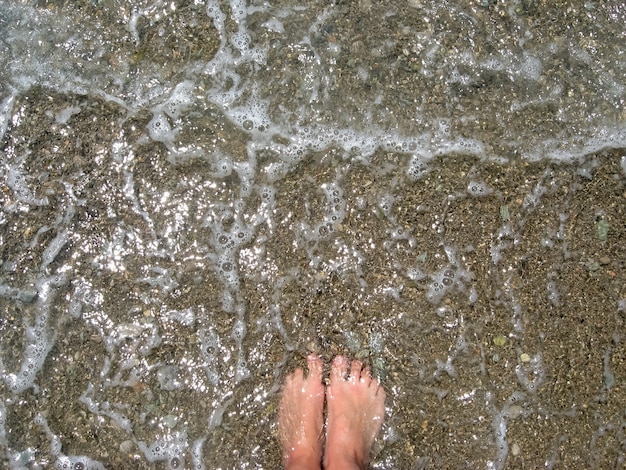 Foto conceito de férias de verão, pés de pés de mulher na água do mar quente e espuma, praia de seixos, descalço, vista superior