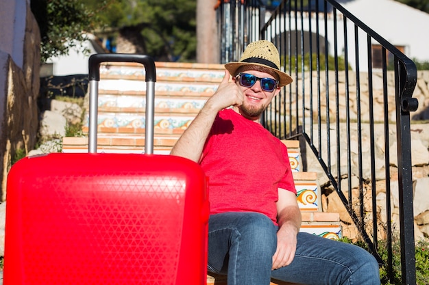 Conceito de férias, viagens e turismo - Homem bonito com mala vermelha sentado em uma escada e mostrando o polegar para cima