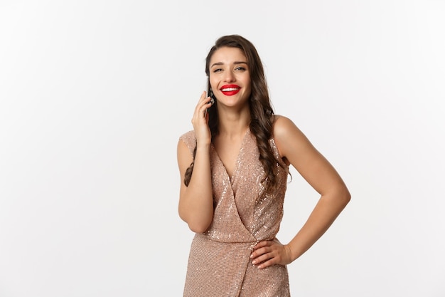 Conceito de festa e celebração de Natal. Mulher jovem e atraente com lábios vermelhos, usando um vestido de luxo, falando no celular e sorrindo, em pé sobre um fundo branco.