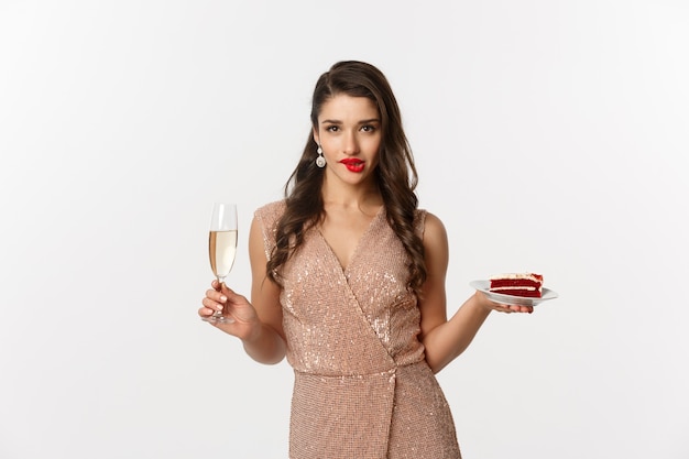 Conceito de festa e celebração. Mulher sexy em um vestido elegante, segurando champanhe e um pedaço de bolo, mordendo o lábio e olhando sedutor para a câmera.