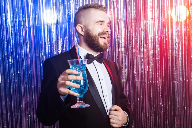 Conceito de festa e feriados do clube - retrato de homem bonito feliz elegante em um terno caro detém coquetel azul sobre fundo brilhante.