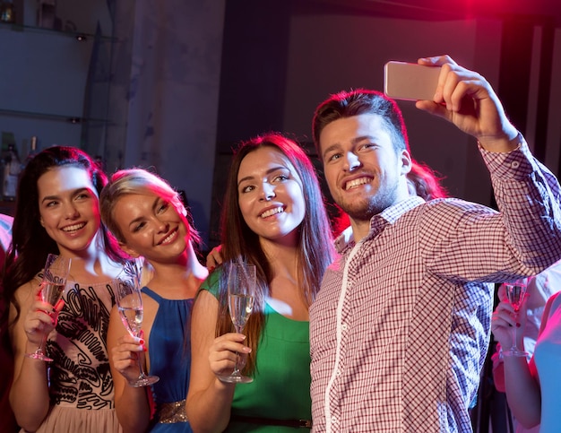 Conceito de festa, feriados, tecnologia, vida noturna e pessoas - amigos sorridentes com copos de champanhe e smartphone tomando selfie no clube