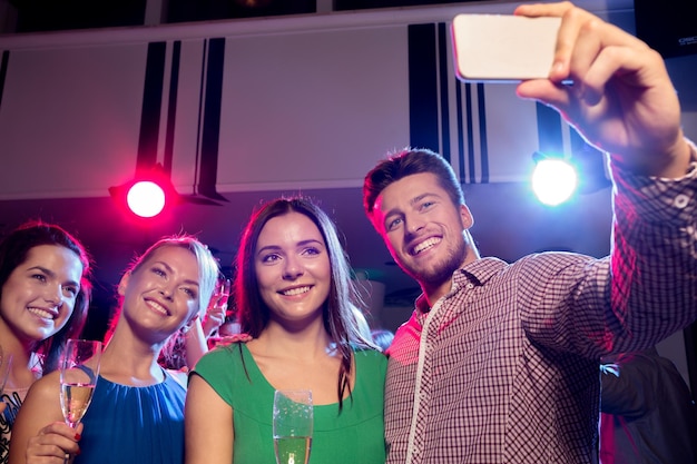 Conceito de festa, feriados, tecnologia, vida noturna e pessoas - amigos sorridentes com copos de champanhe e smartphone tomando selfie no clube