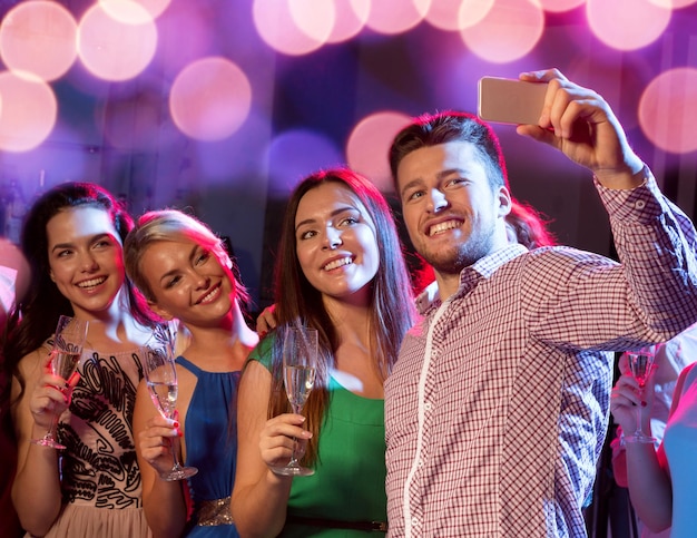 Conceito de festa, feriados, tecnologia, vida noturna e pessoas - amigos sorridentes com copos de champanhe e smartphone tomando selfie no clube