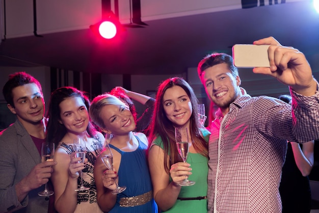 Conceito de festa, feriados, tecnologia, vida noturna e pessoas - amigos sorridentes com copos de champanhe e smartphone tomando selfie no clube