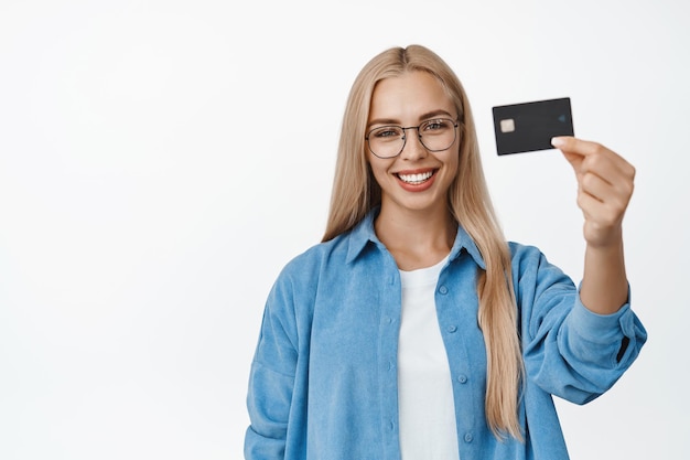 Conceito de finanças e banco Mulher loira sorridente de óculos segurando o cartão de crédito na mão e parecendo feliz recomendando ficar sobre fundo branco