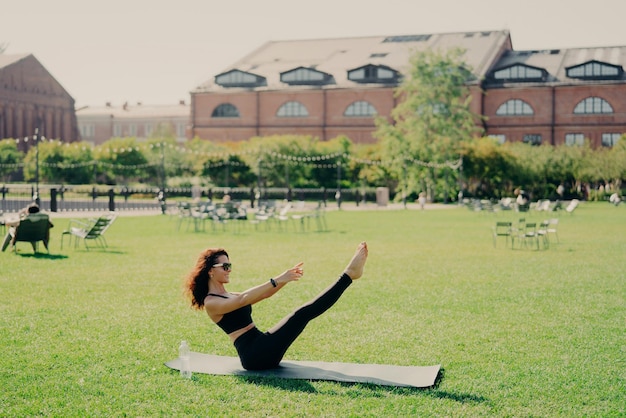 Conceito de fitness e perda de peso de Pilates Jovem morena positiva tem treinamento ao ar livre levanta as pernas poses em karemathas treino regular usa roupas esportivas óculos de sol estica os músculos