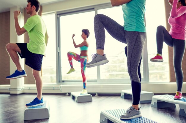 Conceito de fitness, esporte, aeróbica e pessoas - grupo de pessoas sorridentes malhando e levantando as pernas em plataformas de passo no ginásio