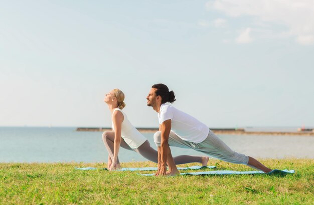 conceito de fitness, esporte, amizade e estilo de vida - casal sorridente fazendo exercícios de ioga em tapetes ao ar livre