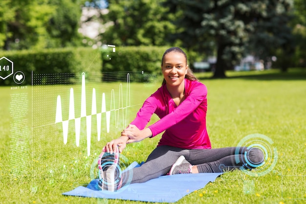 Conceito de fitness, esporte, treinamento, parque e estilo de vida - mulher sorridente esticando a perna no tapete ao ar livre