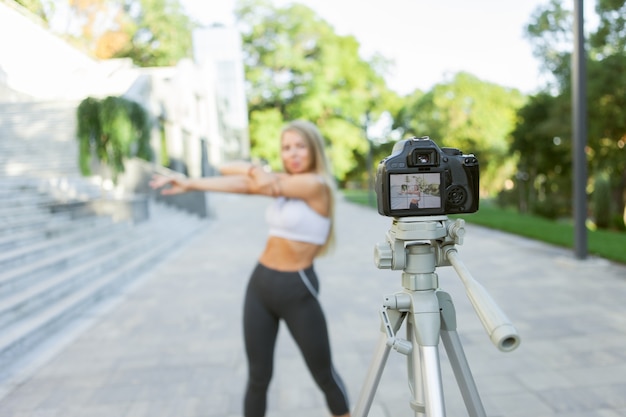 Conceito de fitness, esportes e videoblog. Blogueira de esporte feminino com câmera no tripé, gravando o treinamento de fitness ao ar livre. Foco na câmera