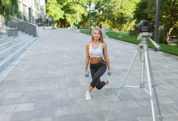 Conceito de fitness, esportes e videoblog. blogueira de esportes femininos com câmera no tripé gravando treinamento físico ao ar livre com halteres