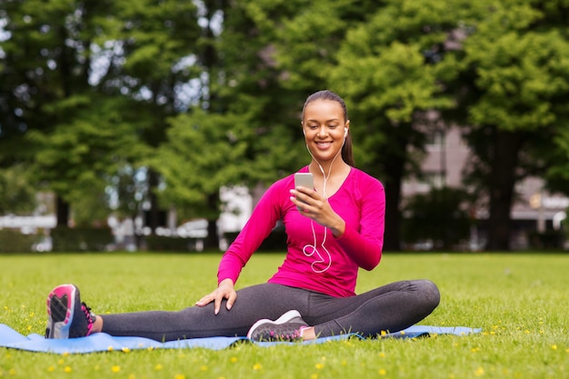 conceito de fitness, parque, tecnologia e esporte - mulher afro-americana sorridente com smartphone e fones de ouvido sentado no tapete ao ar livre