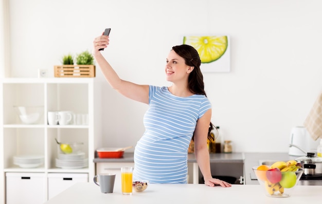 conceito de gravidez, pessoas e tecnologia - mulher grávida feliz com smartphone tomando selfie e tomando café da manhã na cozinha de casa