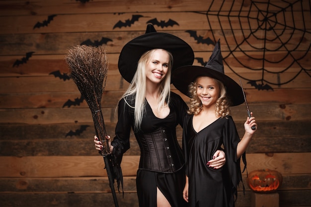 Conceito de Halloween - alegre mãe e filha em fantasias de bruxa, celebrando o Halloween, posando com abóboras curvas sobre morcegos e teia de aranha na parede de madeira.