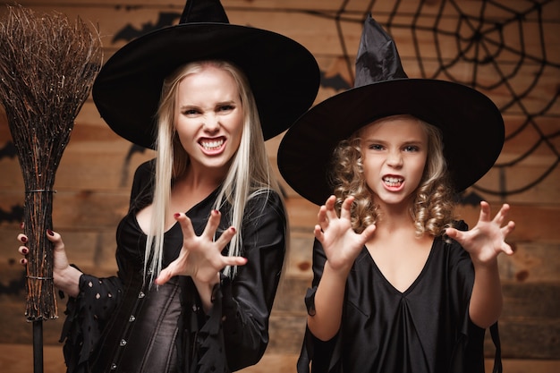 Conceito de Halloween - closeup caucasiana mãe e sua filha em fantasias de bruxa, comemorando o Halloween posando com abóboras curvas sobre morcegos e teia de aranha na parede de madeira.