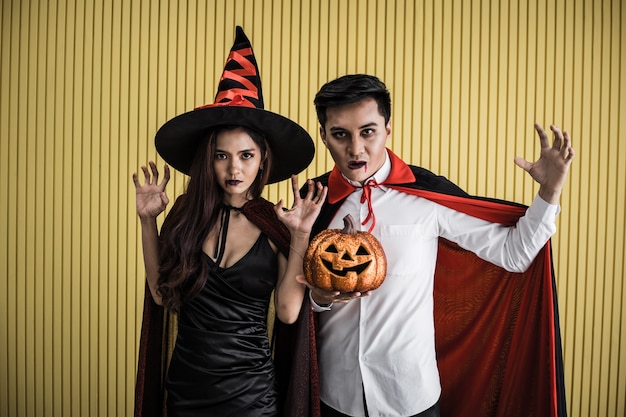 Conceito de halloween de uma jovem mulher asiática na fantasia de bruxa e o homem asiático na fantasia de drácula em fundo amarelo. casal adolescente do retrato vestido de bruxas e drácula para celebrar o festival de halloween.