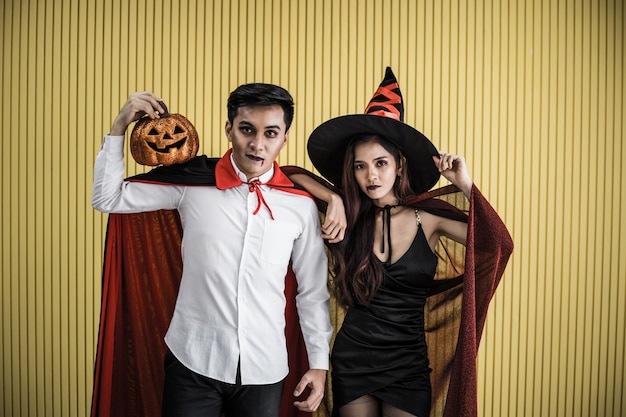 Conceito de halloween de uma jovem mulher asiática na fantasia de bruxa e o homem asiático na fantasia de drácula em fundo amarelo. casal adolescente do retrato vestido de bruxas e drácula para celebrar o festival de halloween.