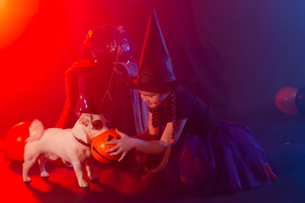 Conceito de Halloween e celebração Garota infantil em fantasia de bruxa com abóbora de Halloween brincando com cachorro jack russell terrier