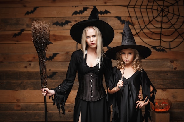 Conceito de halloween - mãe alegre e sua filha em trajes de bruxa comemorando o dia das bruxas posando com abóboras curvas sobre morcegos e teia de aranha no fundo de estúdio de madeira.