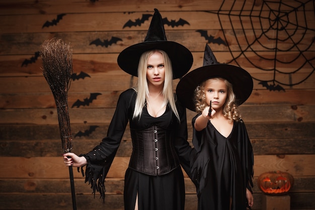 Conceito de Halloween - mãe alegre e sua filha em trajes de bruxa comemorando o Dia das Bruxas posando com abóboras curvas sobre morcegos e teia de aranha no fundo de estúdio de madeira.