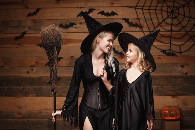 Conceito de Halloween - mãe alegre e sua filha em trajes de bruxa comemorando o Dia das Bruxas posando com abóboras curvas sobre morcegos e teia de aranha no fundo de estúdio de madeira.