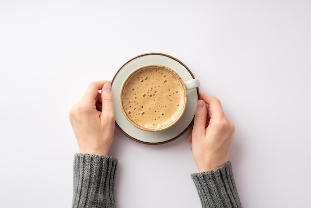 Conceito de humor de outono Foto de primeira pessoa vista superior de mãos femininas em suéter cinza segurando pires com xícara de café espumoso em fundo branco isolado