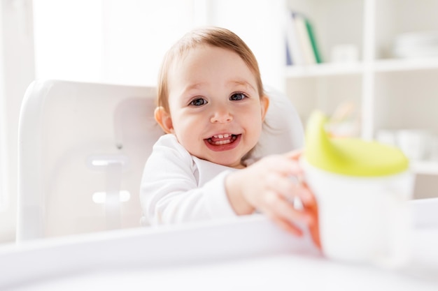 conceito de infância e pessoas - bebê sorridente feliz bebendo do copo de bico sentado na cadeira alta em casa