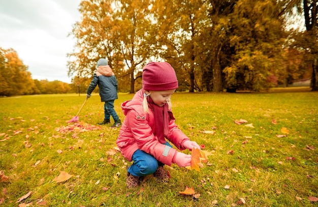 conceito de infância, lazer, amizade e pessoas - menina e menino com rack coletando e trasfegando folhas no parque outono