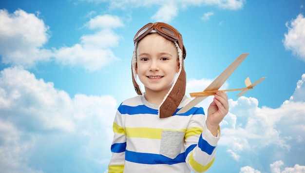 conceito de infância, moda e pessoas - menino sorridente feliz com chapéu de aviador brincando com avião de madeira sobre céu azul e fundo de nuvens