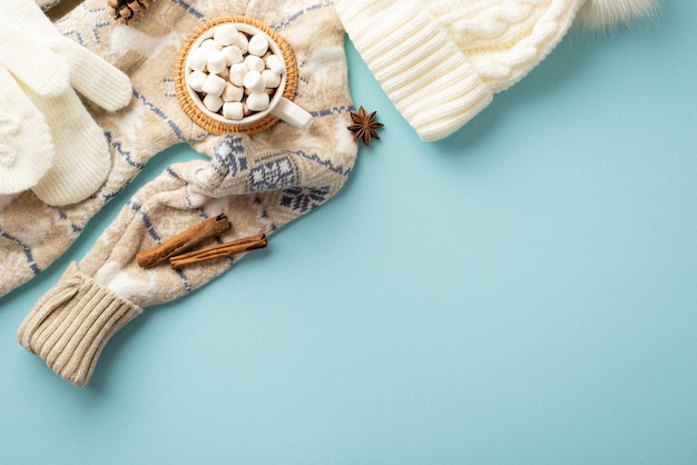 Conceito de inverno Foto de vista superior de luvas de chapéu de malha aconchegante caneca de chocolate quente com marshmallow em jogo americano de vime anis e paus de canela em fundo azul pastel isolado com copyspace