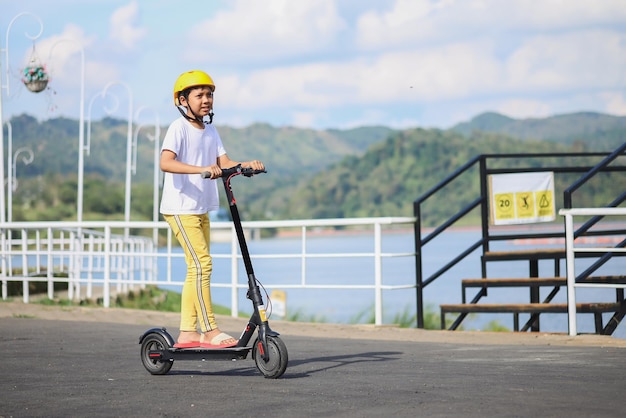 Conceito de lazer da juventude. Retrato de corpo inteiro de menino animado andando de e-scooter preto no parque do rio.