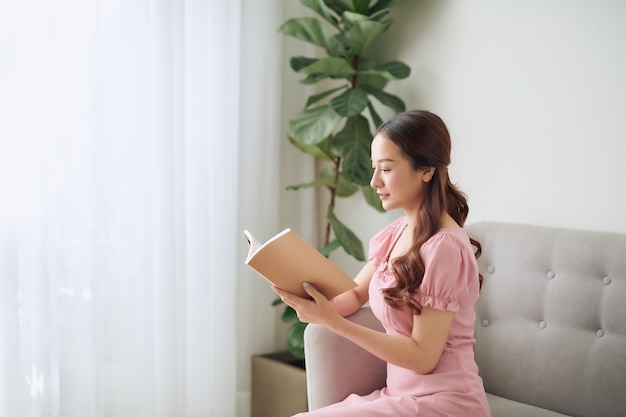 Conceito de lazer, literatura e pessoas - sorrindo mulher asiática lendo livro em casa