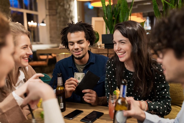 conceito de lazer, pessoas e férias - amigos felizes com bebidas, dinheiro e smartphones no bar ou café