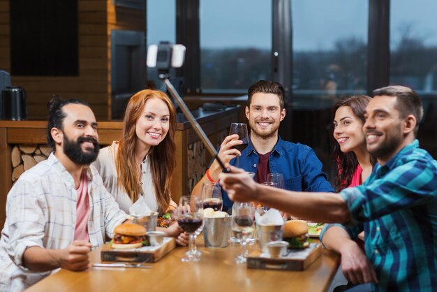 conceito de lazer, tecnologia, amizade, pessoas e feriados - amigos felizes jantando e tirando fotos com o bastão de selfie do smartphone no restaurante