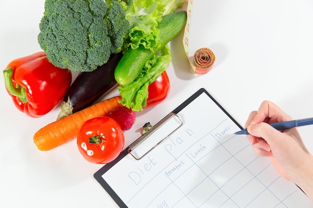 Conceito de local de trabalho do médico nutricionista. Pessoa do sexo feminino com as mãos escrevendo plano de dieta, composição de vegetais frescos, fita métrica e telefone celular isolado no fundo branco, vista superior