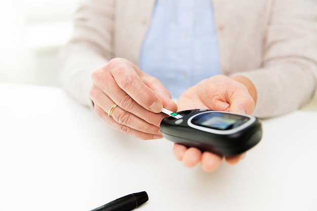 Conceito de medicina, idade, diabetes, saúde e pessoas - close-up de uma mulher idosa com glicosímetro e faixa de teste, verificando o nível de açúcar no sangue em casa