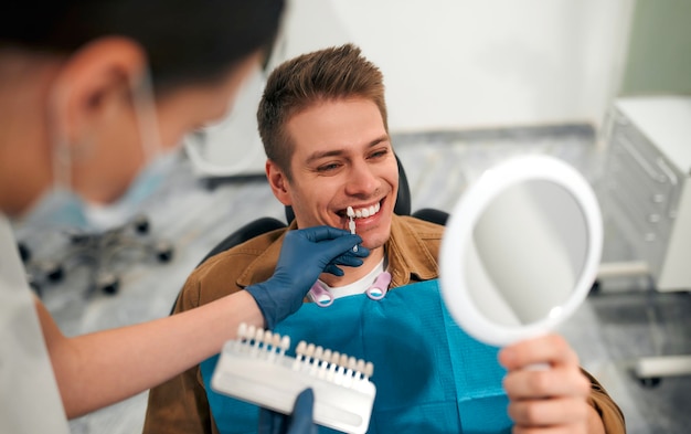 Foto conceito de medicina, odontologia e saúde - closeup de um dentista com amostras de cor de dente, escolhendo uma tonalidade para os dentes de um paciente do sexo masculino em uma clínica odontológica, olhando para um espelho.