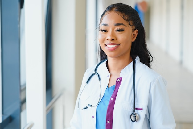 Conceito de medicina, profissão e saúde - feliz e sorridente médica afro-americana de jaleco branco com estetoscópio sobre fundo de hospital