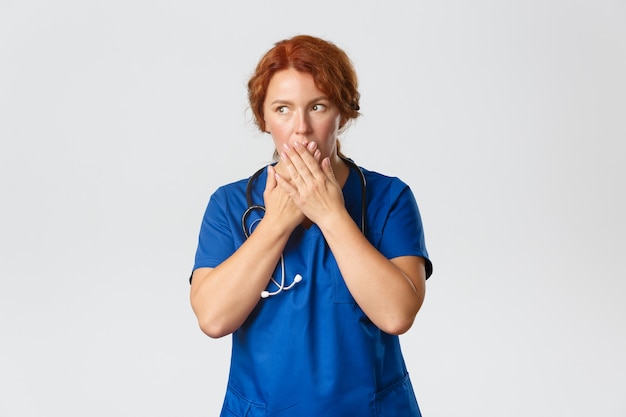 Conceito de medicina, saúde e coronavírus. Retrato de uma médica ruiva chocada e impressionada, enfermeira ouve fofoca no hospital, cobre a boca com a mão e desvia o olhar atônito com a notícia.