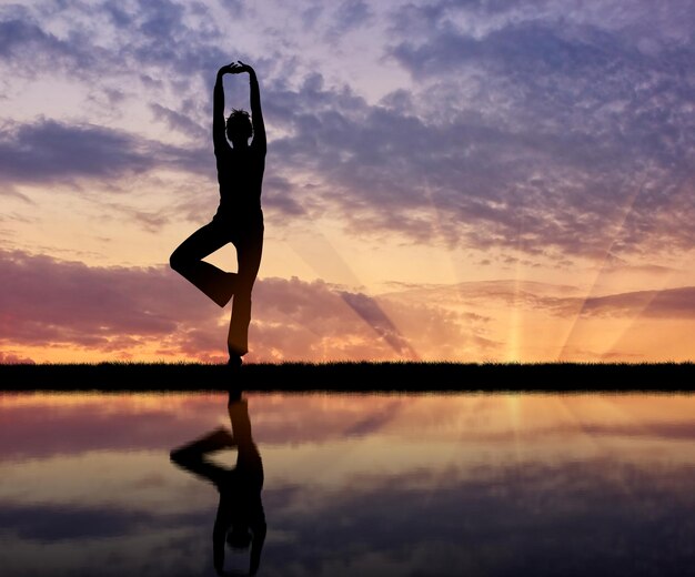 Conceito de meditação e relaxamento. Silhueta de uma garota praticando ioga ao pôr do sol e reflexo na água