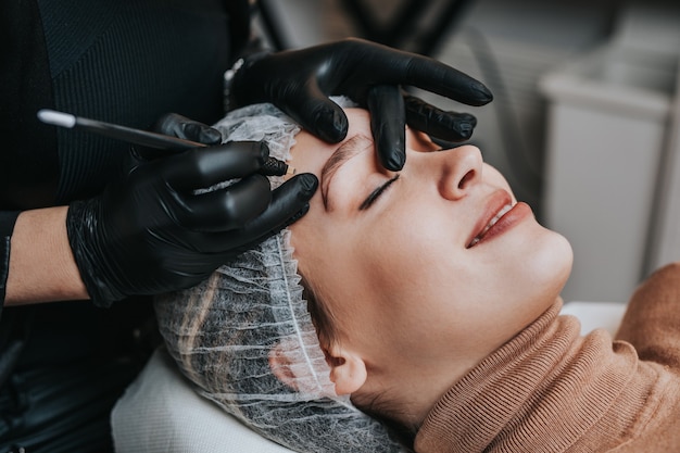 Conceito de microblading de sobrancelhas, procedimento de maquiagem definitiva de sobrancelha. A especialista em beleza está usando escudo e máscara protetora devido à pandemia do Coronavírus.