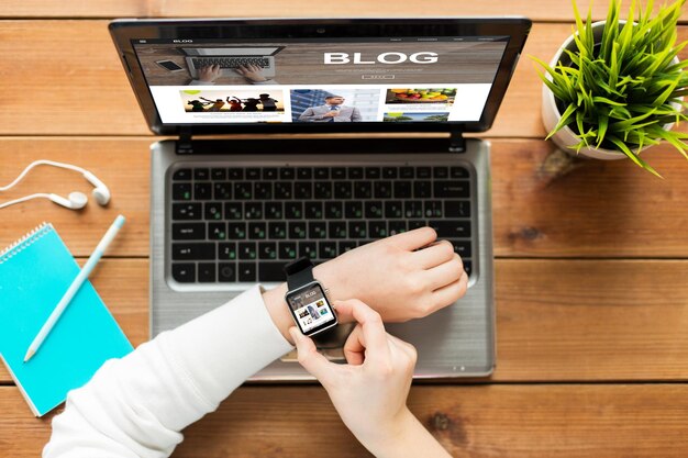 Foto conceito de mídia, internet, negócios, pessoas e tecnologia - close-up da mulher com relógio inteligente e laptop com página da web do blog na tela na mesa de madeira